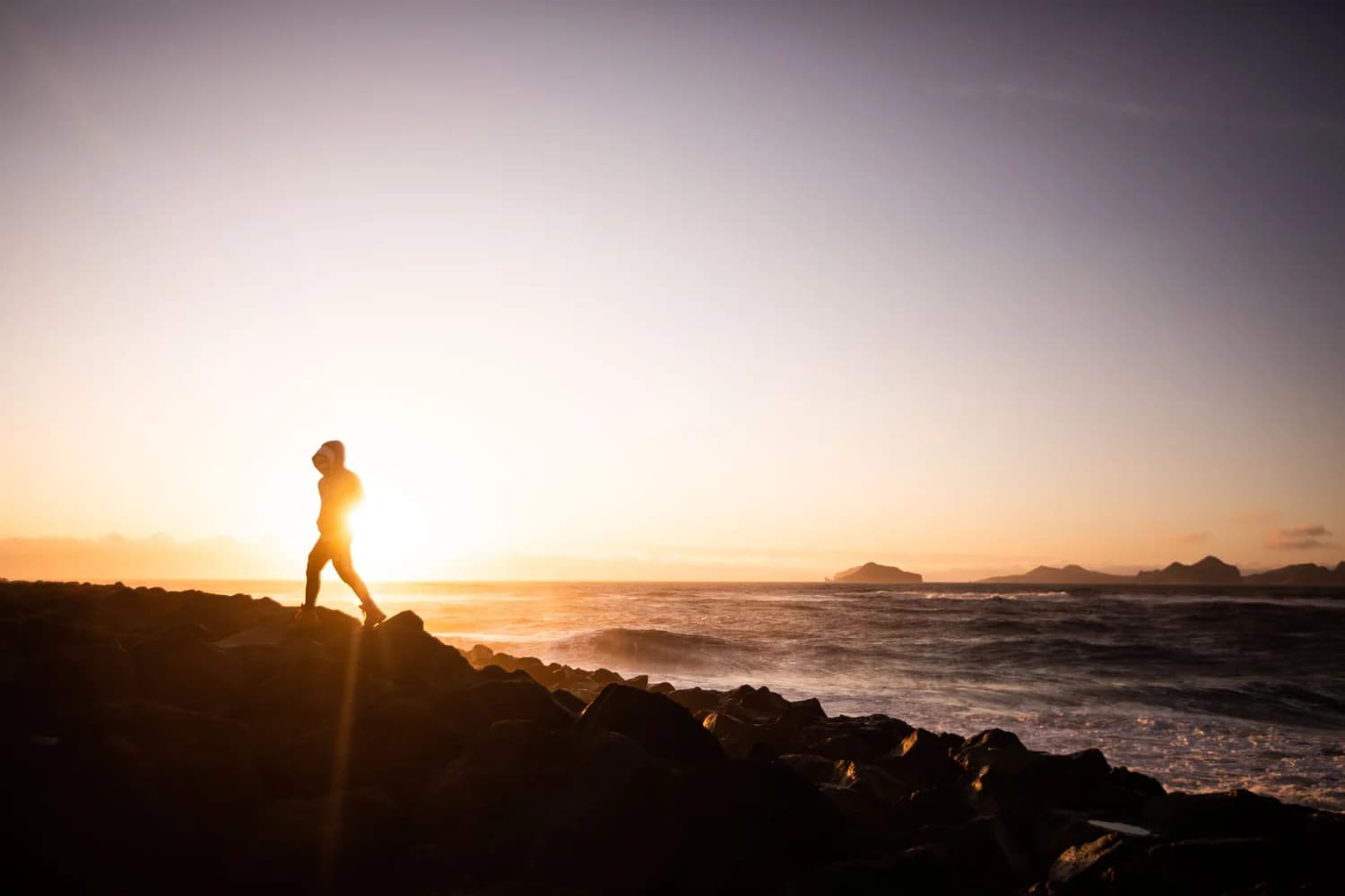 A lone figure walking across coastal rocks as the sun sets over the ocean